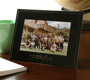 Glossy black cardboard frame sits on a dark wood table surrounded by a notebook, coffee cup, and globe. Gold foil imprint on the frame reads, Be the spark that inspires the world.