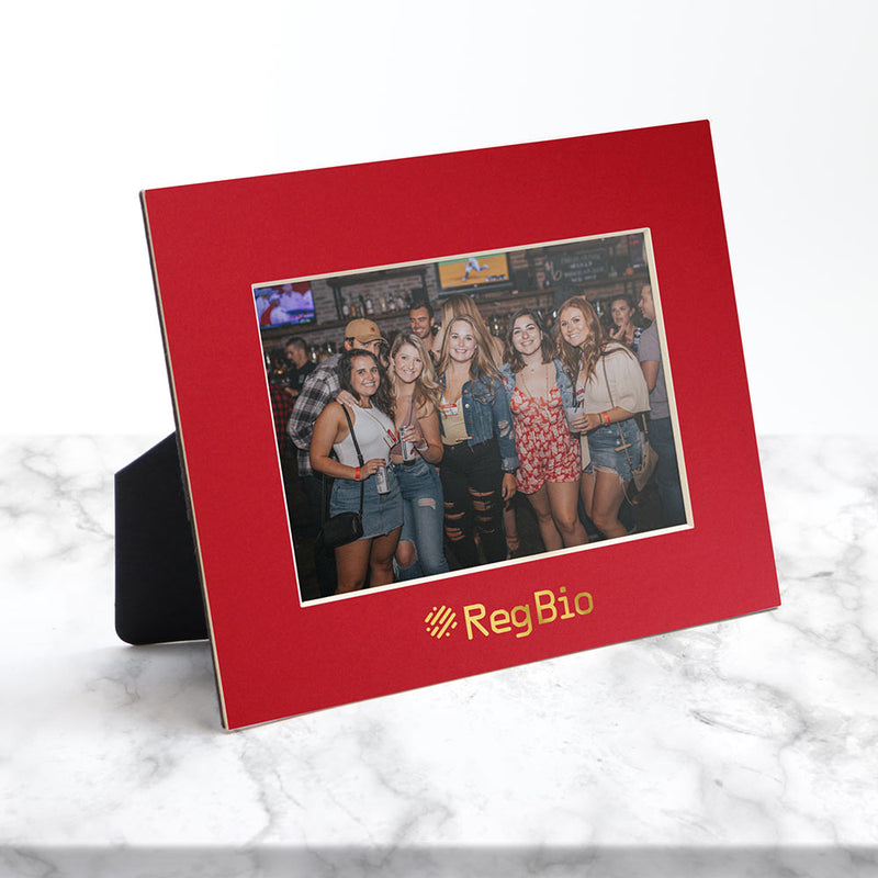 Horizontal red frame sits on a white marble counter and has a gold foil logo imprint.