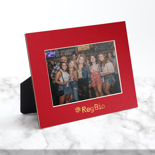 Horizontal red frame sits on a white marble counter and has a gold foil logo imprint.