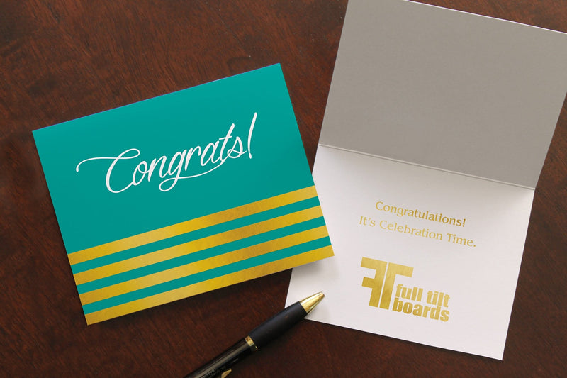 Tela congrats card lies on a wood table. One card is closed, the other open, showing a congratulations sentiment and company logo stamped in gold foil.