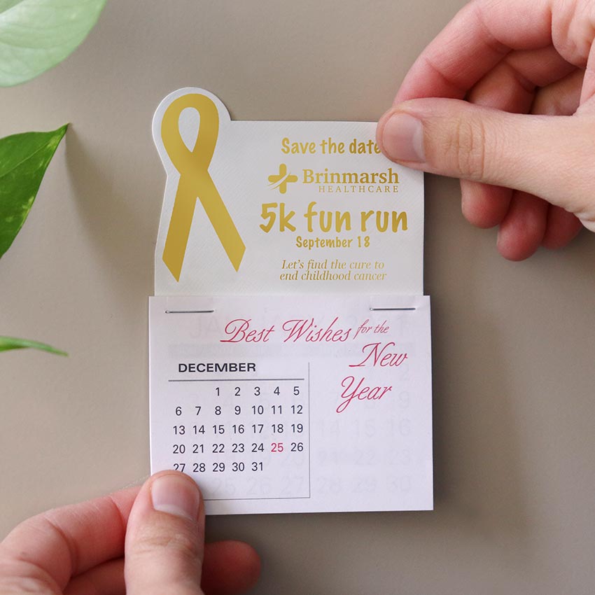 Someone is sticking a small white calendar to a cabinet. The calendar has a gold imprint with a ribbon and info for a fundraiser for a healthcare organization.