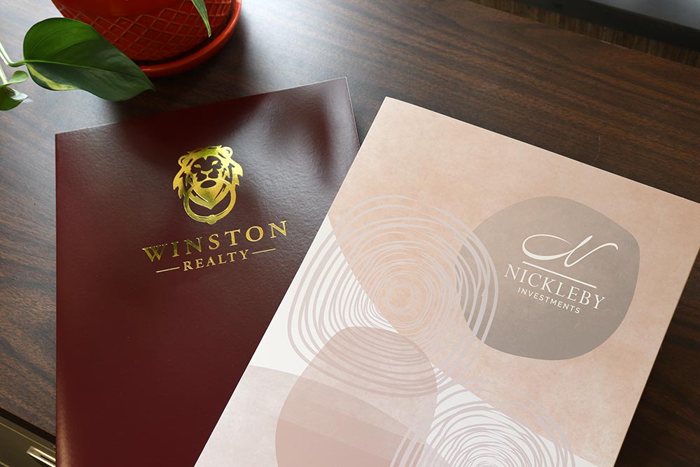 Two pocket folders lie on a wooden cabinet. One is burgundy with a gold foil imprint, the other is white but is printed with a company's brand design.