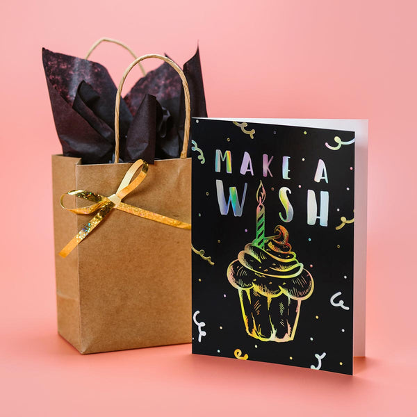 Black greeting card with gold and silver foil birthday cupcake design stands next to a kraft paper gift bag with gold ribbon and black tissue paper.