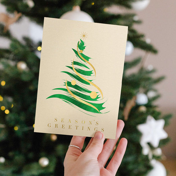 Someone holds up the brushstroke tree card in front of a lit, decorated Christmas tree.
