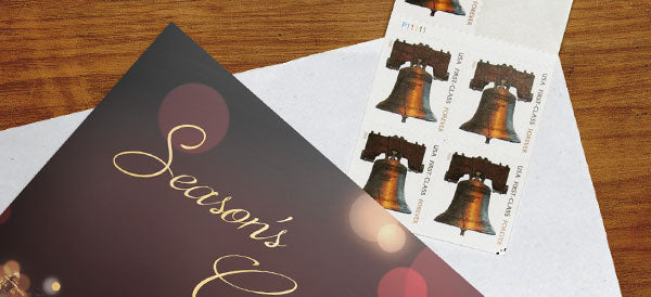 A holiday greeting card lies on a white envelope with first-class USPS stamps next to it.