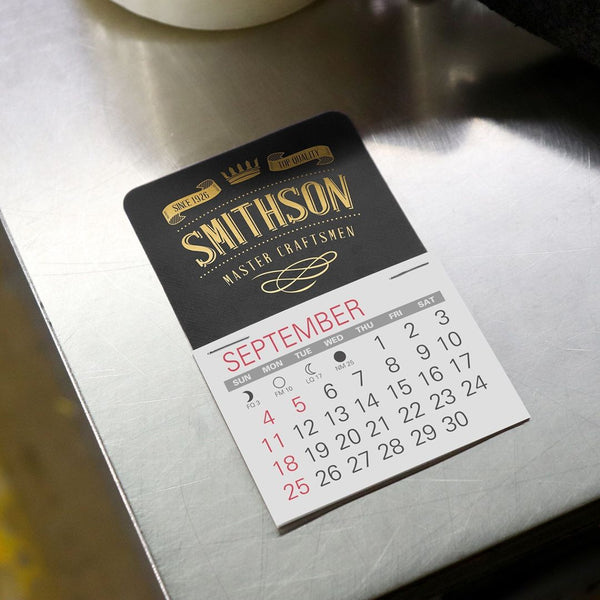 Black calendar with gold foil craftsmen company logo imprint sticks to a metal table.