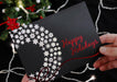 Someone holds a black Christmas card with a silver snowflake wreath and red foil Happy Holidays design