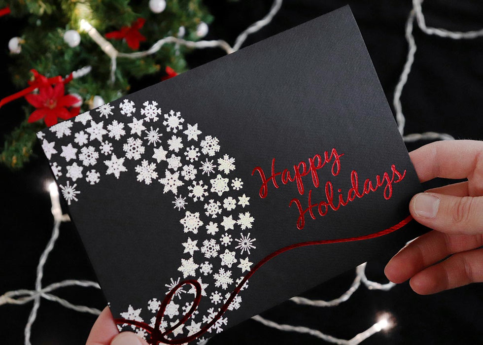 Someone holds a black Christmas card with a silver snowflake wreath and red foil Happy Holidays design