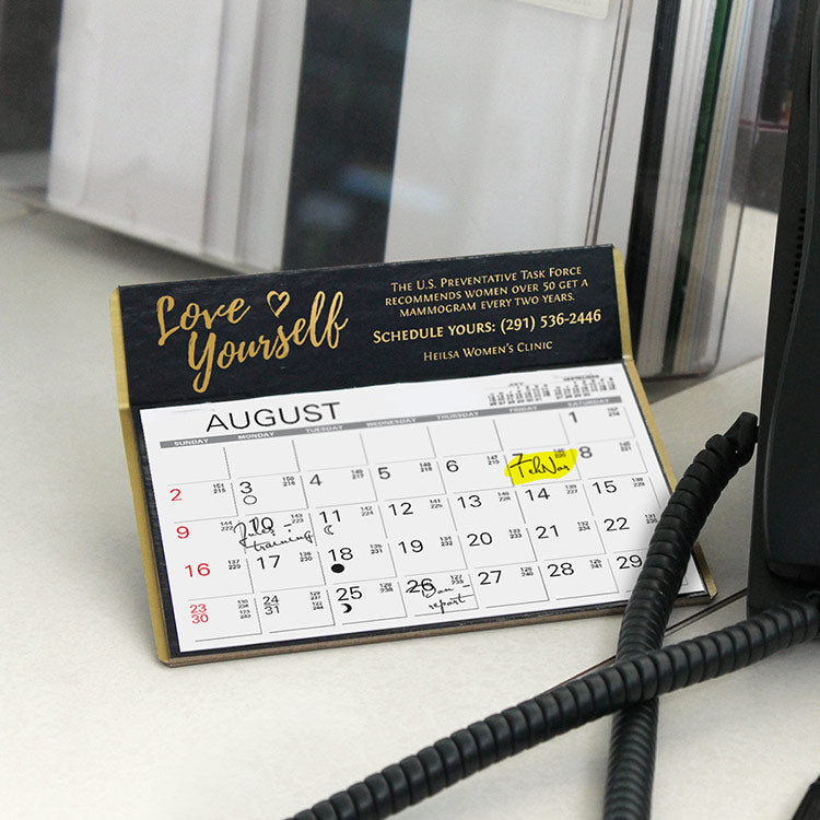 Black calendar with gold sides and golf foil logo imprint sits on an office desk next to a black phone.