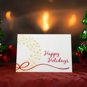 The ivory Christmas card with gold wreath design sits on a red table cloth in front of a lit fireplace. The card is flanked on both sides with small tinsel Christmas trees.