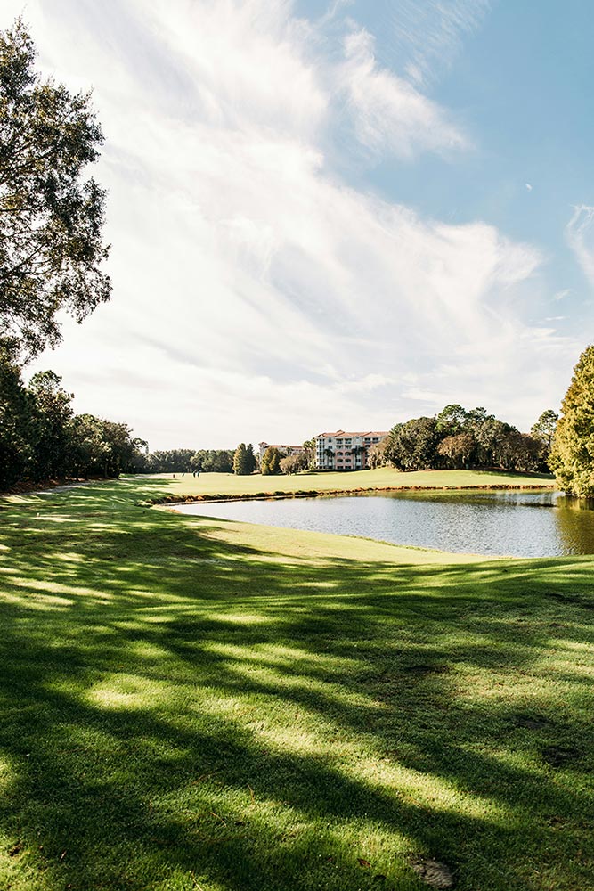 Golf course green with dappled sunlight in the foreground, a pond to the left, and a sunlit club house in the background against a sunny sky with some wispy clouds.