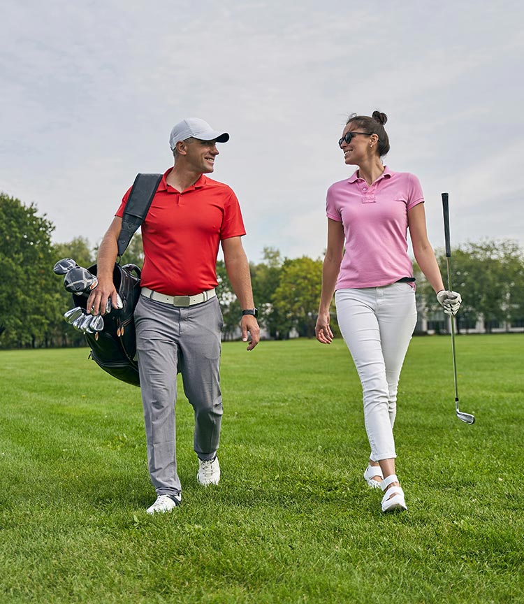 A man and woman walk toward the camera on the golf course, smiling at each other. The woman is carrying a club, the man is carrying the bag of clubs.
