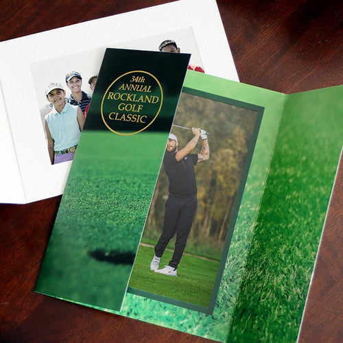 A green golf gatefold photo folder with gold foil event logo imprint on one flap lies on a white photo folder that's dimpled like a golf ball.