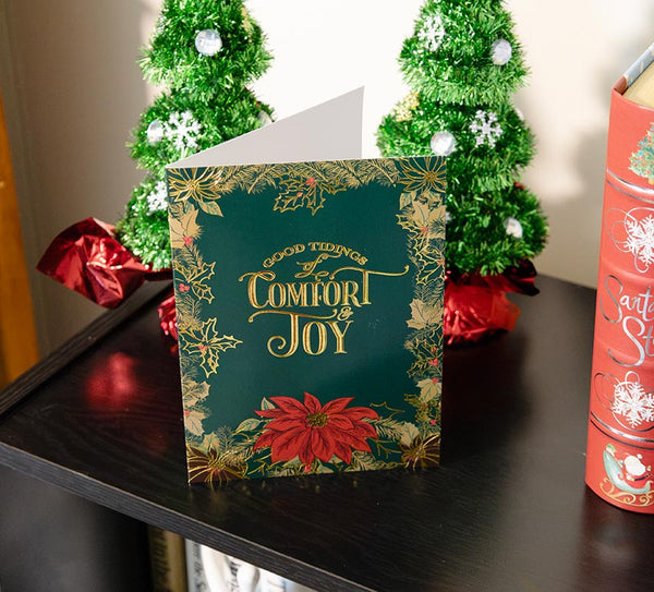 Green, gold, and red poinsettia holiday card sits on a wooden shelf, surrounded by Christmas books and small tinsel trees.