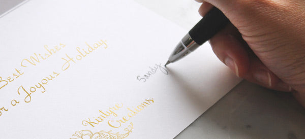 Someone writes a message in a greeting card that has a gold foil stamped sentiment and logo imprint.