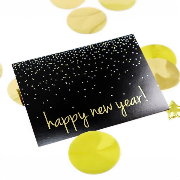 Black new years card with gold and silver foil confetti design. Sits on a bright white surface with large gold confetti dots.