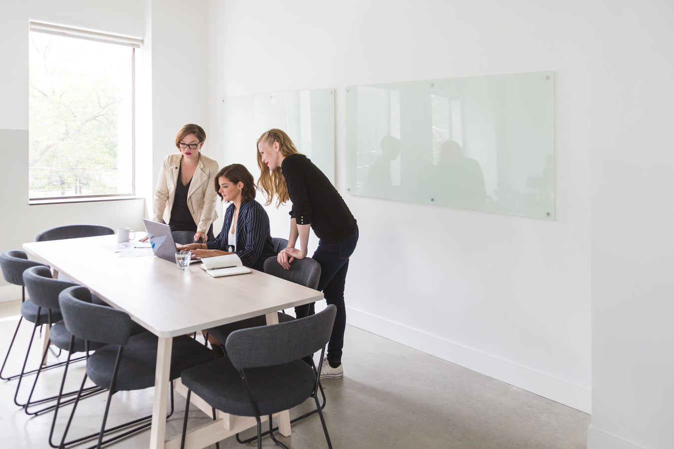 Three woman look at a laptop sitting on a light wood table in a bright, sunlit conference room.