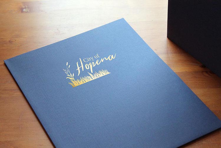 Navy blue pocket folder lays on a wooden table with a gold foil logo imprint for a city.