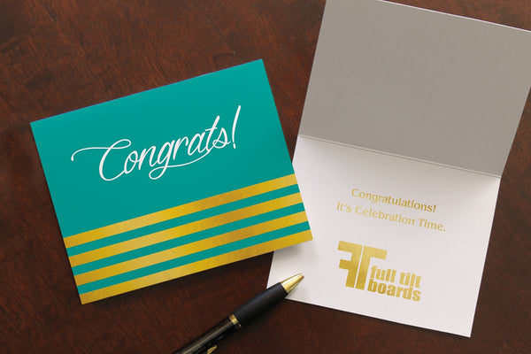 Tela congrats card lies on a wood table. One card is closed, the other open, showing a congratulations sentiment and company logo stamped in gold foil.