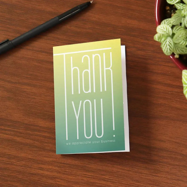 Gradient thank you note card lies on a wooden desk, surrounded by a potted plant and black pen.