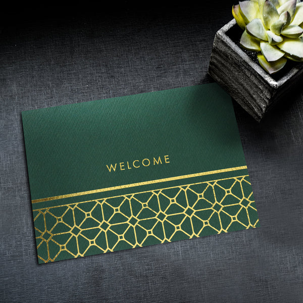 Green greeting card with gold foil lattice and welcome design lies on a black textured surface next to a small potted succulent plant.