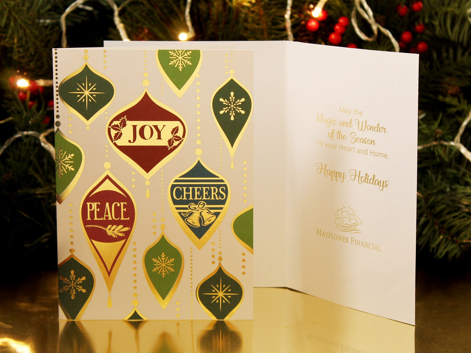 Cream-colored Christmas card stands on a golden surface in front of decorated evergreen boughs. The card has a red, green, and gold vintage ornament design with the words Peace, Joy, and Cheers. Inside the card is a holiday sentiment stamped with a company logo.